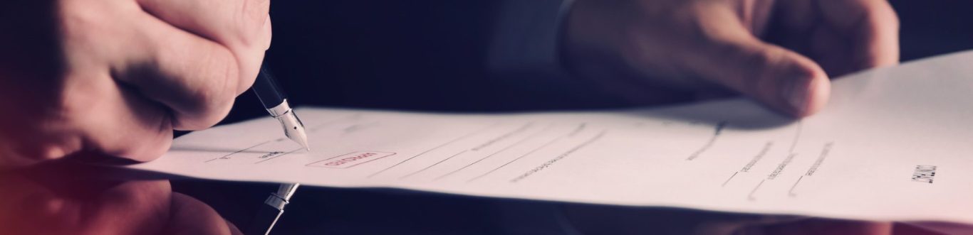 firma documenti Man’s hand signing a document on a desk with a red surface.